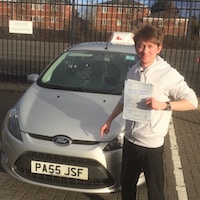 Owen Parker proudly holding his Practical Driving Test pass certificate next to the Driving School car.