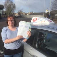 Gill Harris – Jim #3 Gill Harris smiling and holding her practical driving test pass certificate standing next to the JSF Driving School car