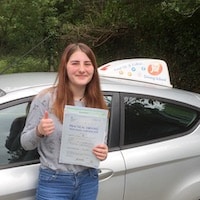 New driver Megan Lambert smiling next to the JSF Driving School car with her practical test pass certificate and thumbs up