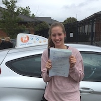Smiling new driver Claire Hughes holding her certificate after passing her driving test first time, standing next to the JSF Driving School car at Gloucester Test Centre