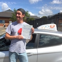 Smiling new driver Sam Daykin holding his certificate after passing his driving test first time, standing next to the JSF Driving School car at Gloucester Test Centre